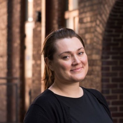 A woman in a black top and brown hair pulled back smiles broadly at the camera with a brick laneway out of focus in the background.